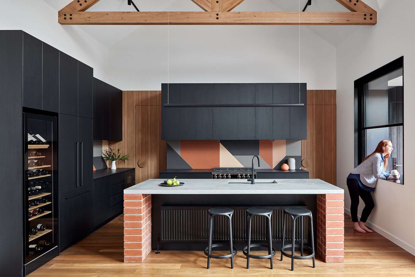 A red brick and concrete island acts as a centerpiece in the kitchen. The benches to the rear and side of island are made from Dekton Sirius reconstituted stone and are surrounded by black laminate cabinetry. The tiles used for the backsplash are Tierras by Patricia Urquiola for Mutina.