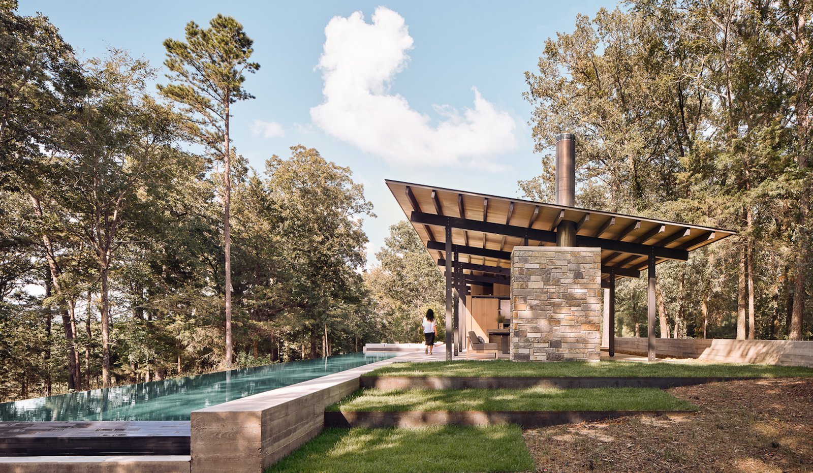 Aegean Pool House’s expansive roof structure shelters the indoor/outdoor living spaces beneath it. A freestanding stone fireplace anchors the west side of the living and dining area.