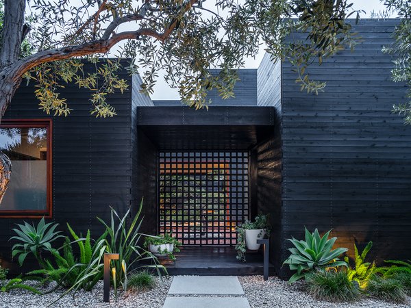 A screen divides an entry porch from the first of several private courtyards and outdoor areas. The home is clad in ebony-stained cedar siding, which contrasts with mahogany casework. 