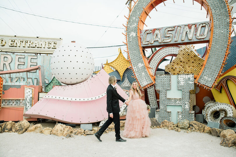 two light-skinned people in formal dress walk through a dumping ground for old neon signs