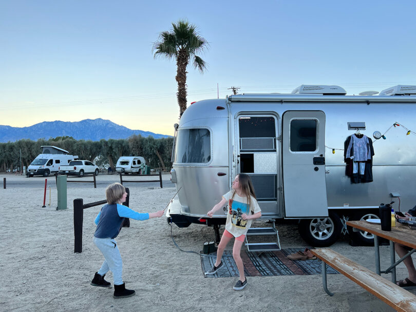 two children play outside of an airstream with palm trees and mountains in the background