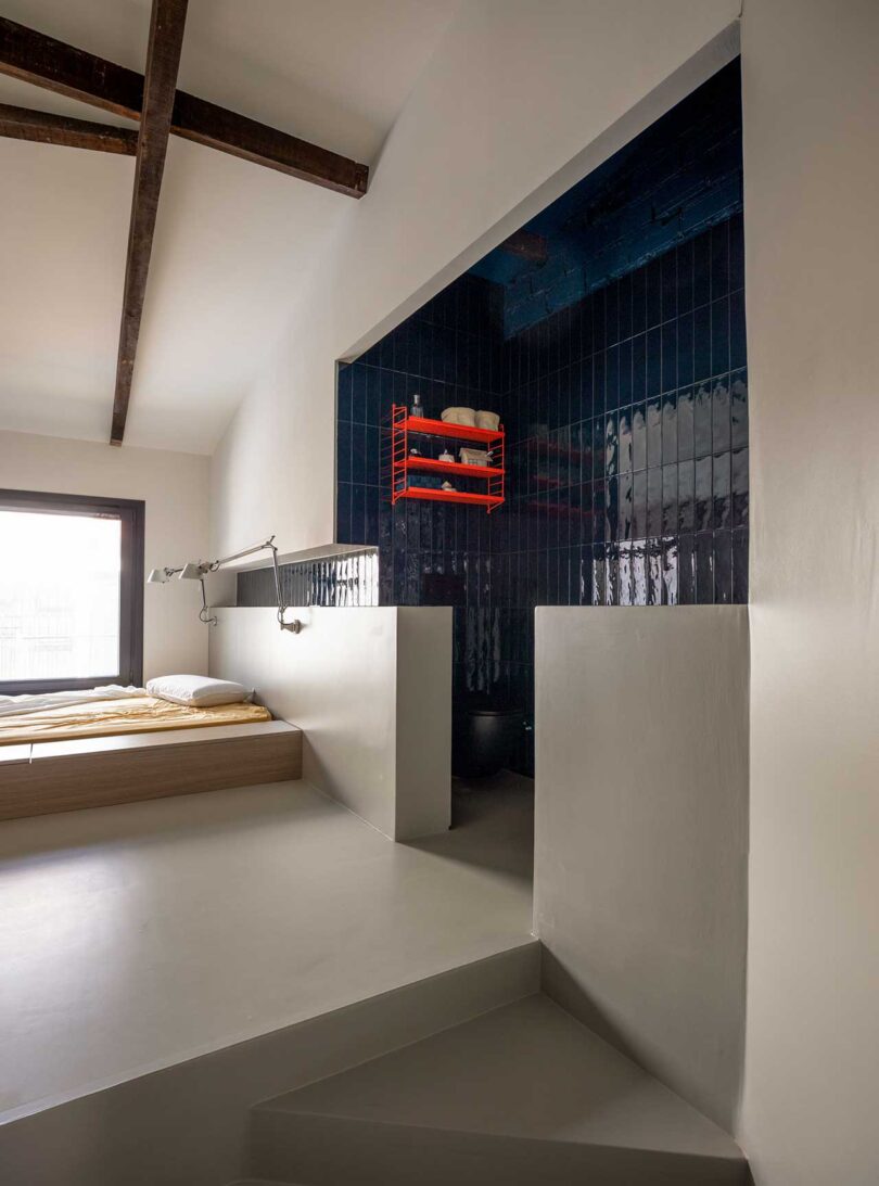 view into open bathroom in modern bedroom with dark blue glossy tile and red shelf