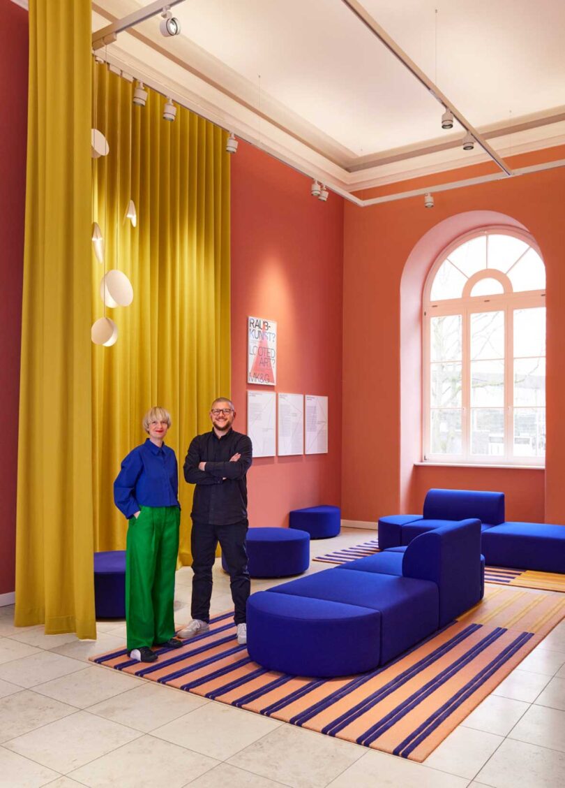 two people standing in a large pink room with modular blue sofa and gold curtains