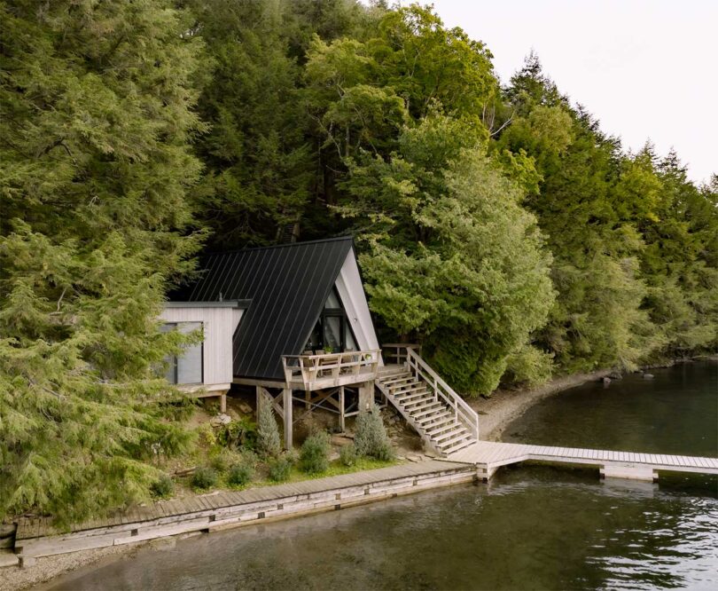 Angled exterior view from lake looking to black a-frame elevated cabin jutting out from trees