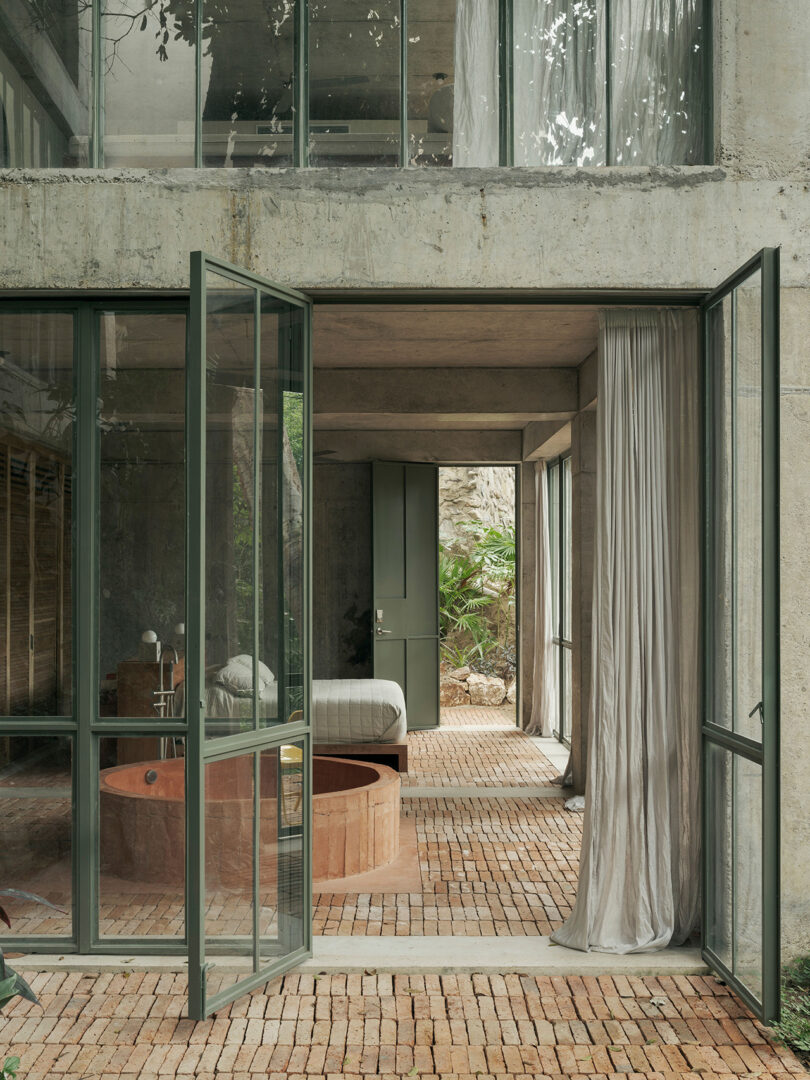 Outside looking into NICO Sayulita's Jungle suite, with paned glass window doors open revealing a circular concrete tub and bed in the background.