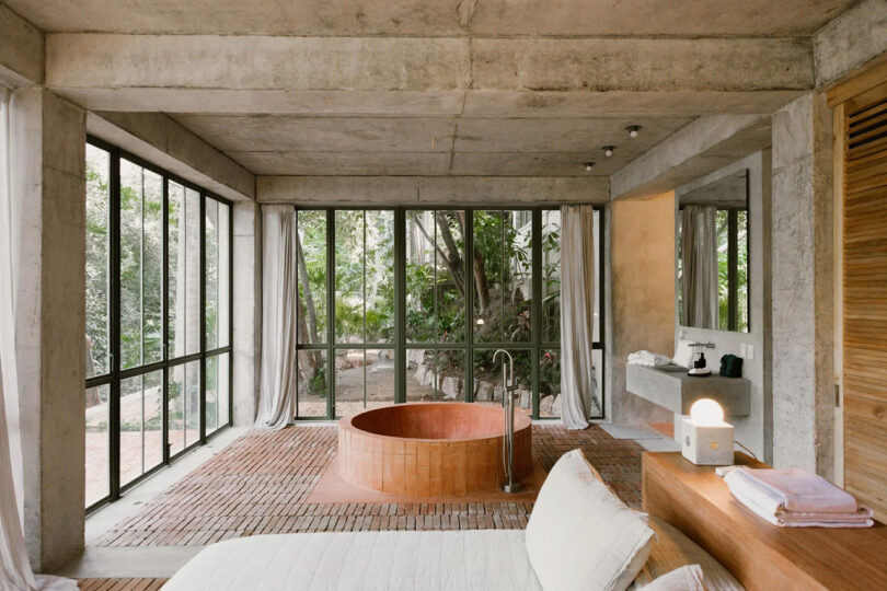 Interior shot of NICO Sayulita's Jungle suite, with paned glass window doors open revealing a circular concrete tub and bed in the background, side of king sized bed in foreground.