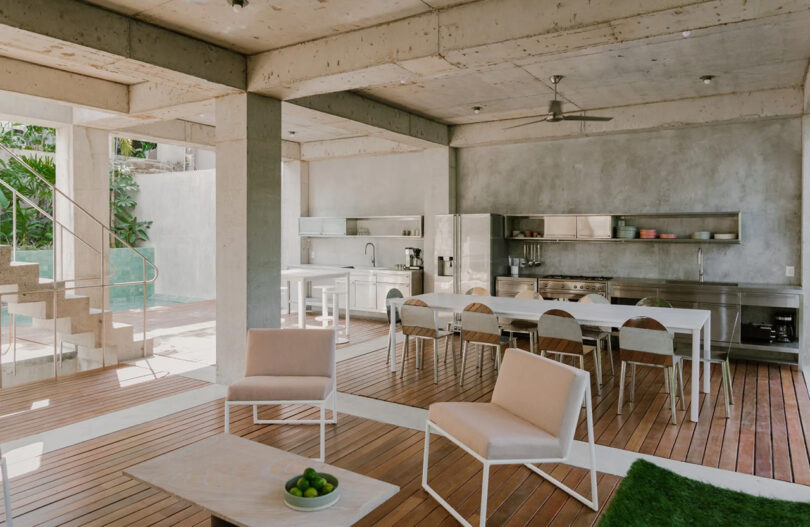 An open air kitchen with eight dining chairs and dining table in white over wood slat flooring inside a concrete clad interior. Two padded patio chairs and low coffee table in the foreground with ceiling fan in background.