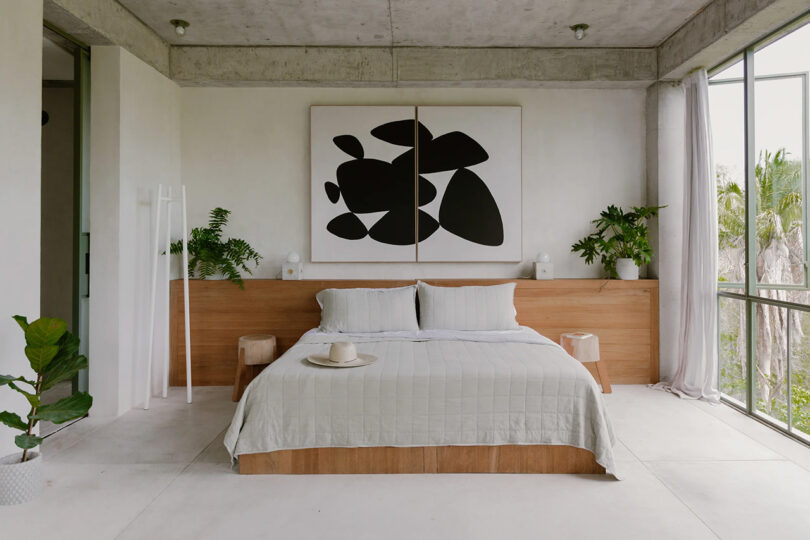 Wood platform kingsize bed in center of white bedroom with two panel abstract artwork over the bed covered in white bedding, surrounded by potted tropical plants and large floor to ceiling windows to the left revealing a tropical setting.