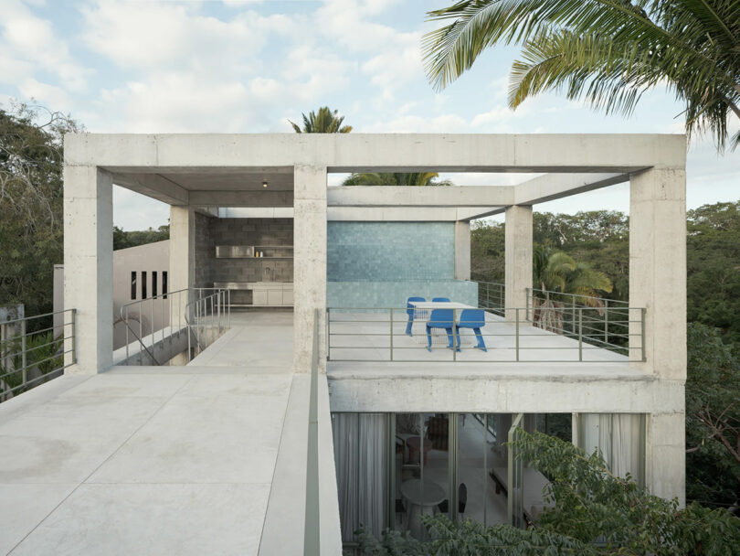 Exterior shot of concrete beam framed terrace with four blue outdoor chairs and table, with turquoise rooftop plunge pool in background to the right. Palm trees surrounding front and back.