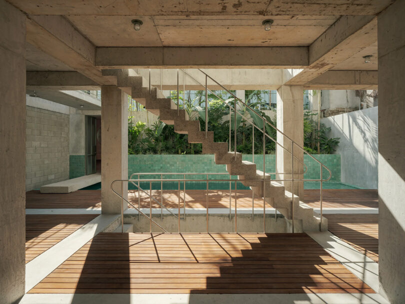 Floor level with floating staircase in the center casting a shadow across open air concrete interior, with turquoise tile lined lap pool in the background.