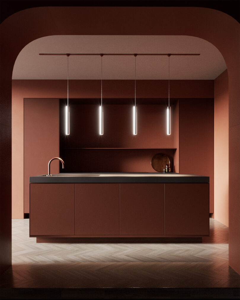 Four cylinder pendant lamps hanging over a modern kitchen island through an arched doorway. Kitchen is painted a clay red with herringbone wood flooring.