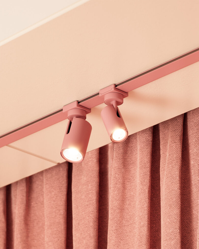 Two pink architectural track heads pointing in opposite directions installed onto ceiling with pink fabric pleated curtain in similar color in background.