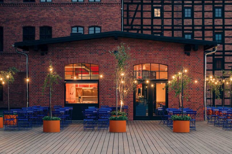 evening view of large brick industrial building with cafe on lower level with outdoor space filled with blue chairs and tables