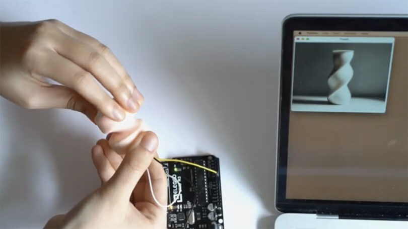 Person's hands using Promptac connected to a rubbery knob piece being twisted to manipulate and twist an AI generated image of a white vase on a nearby laptop screen.