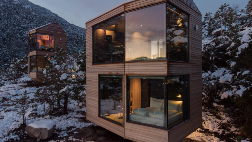 A majestic panoramic view of the nearby fjord located below the series of cabins from the glass facade covered bedroom. Interior is furnished with armchair, bed and pouf.