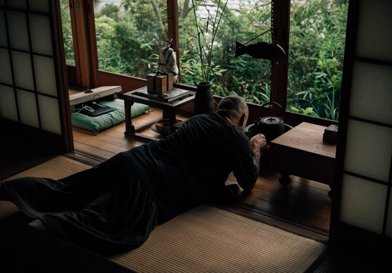 MAKHNO Studio's Serhii Makhno on his stomach across a tatami floor interacting with a traditional Japanese hanging teapot, with garden views in the background. 