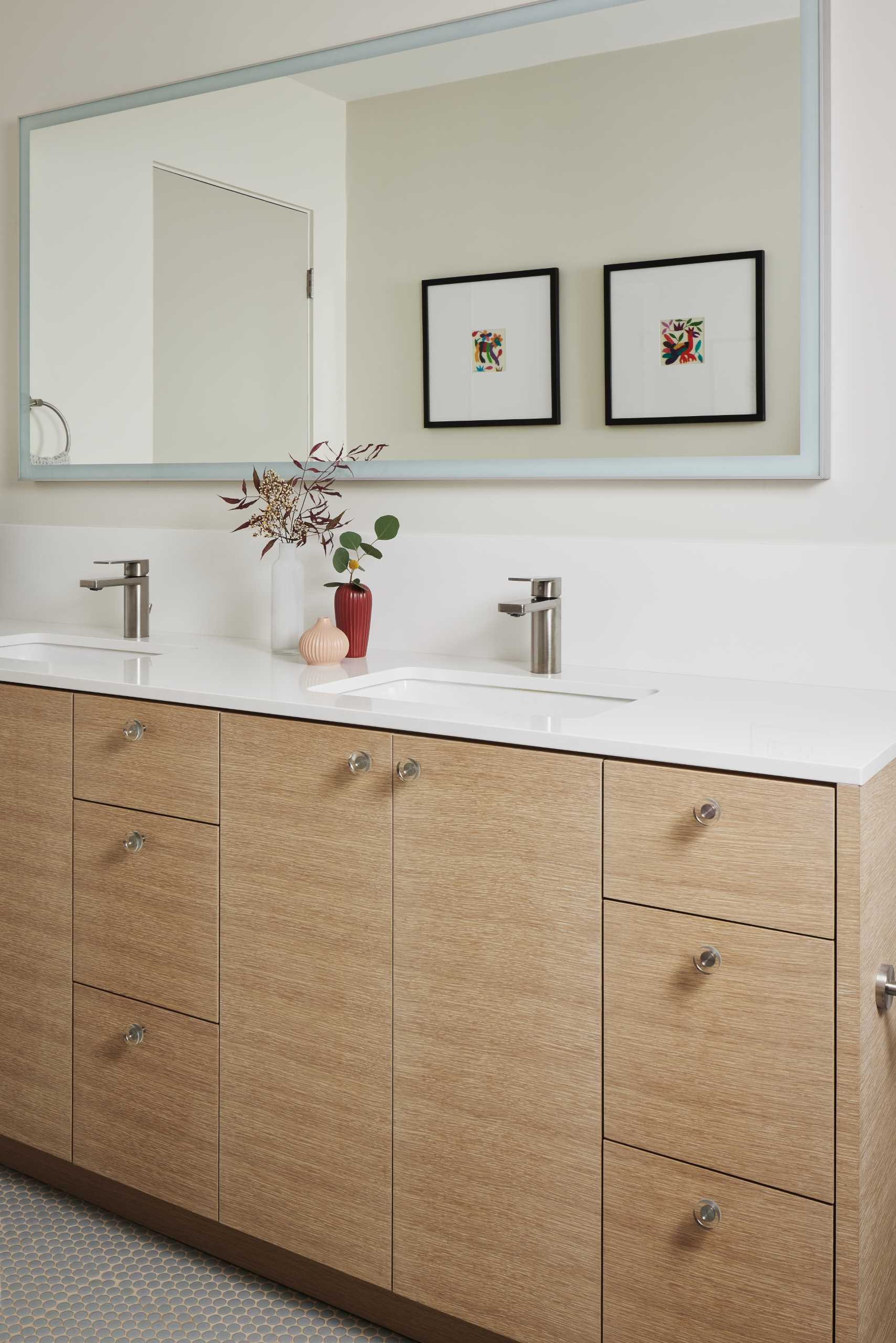 The updated bathroom is bright and open and now includes a double vanity with undermount sinks, a long horizontal mirror, and tile that wraps from the end of the vanity and around the wall to the shower.