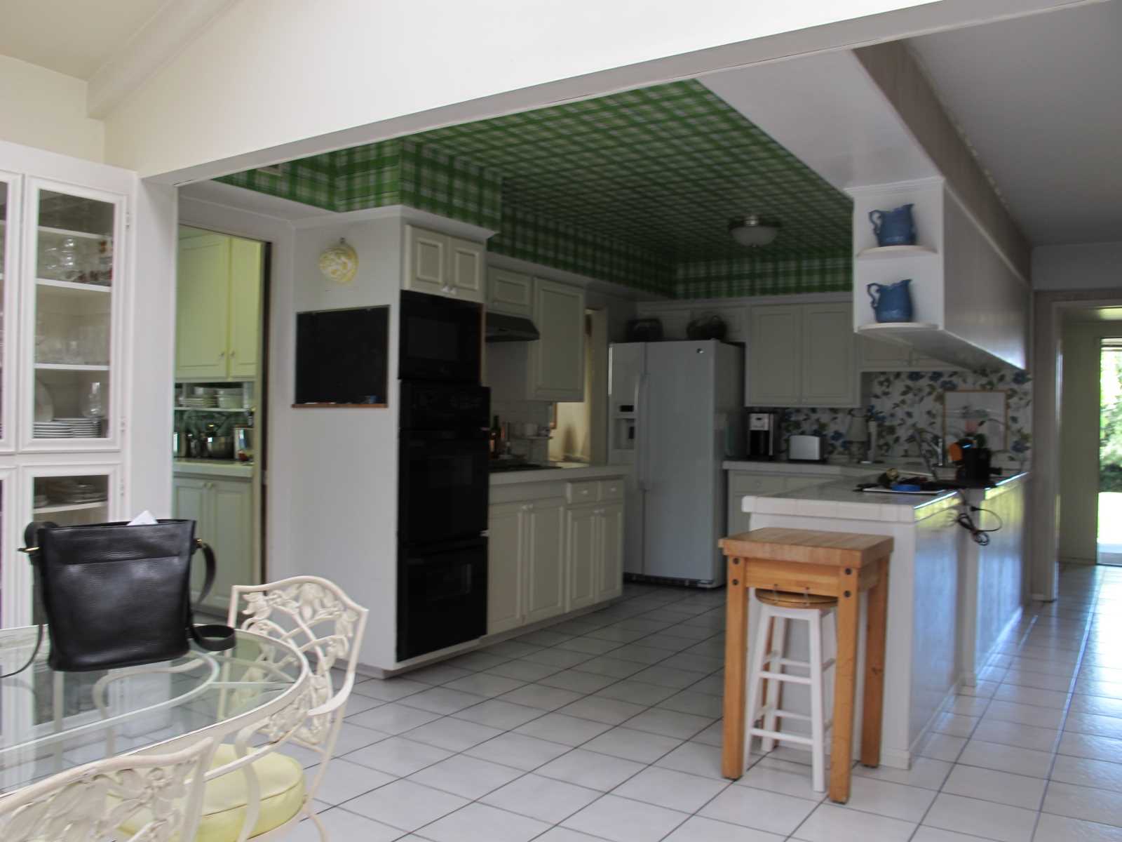 Before - The original kitchen had a tiled floor and countertops, with white cabinets, mismatched appliances, a green tartan wallpapered ceiling, and a floral wallpaper backsplash.