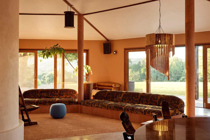 Large length of vintage sofa seating in a the Sun Ranch lobby room with warm yellow hued walls, vaulted ceilings, a small blue ottoman and wooden side chair. A wood dining table can be seen in the foreground alongside a beaded spiral chandelier, with a green tree linked landscape and lawn in the background visible through large patio windows.