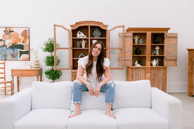 light-skinned woman with long dark hair sits on the back of a white couch with two wood armoires behind her