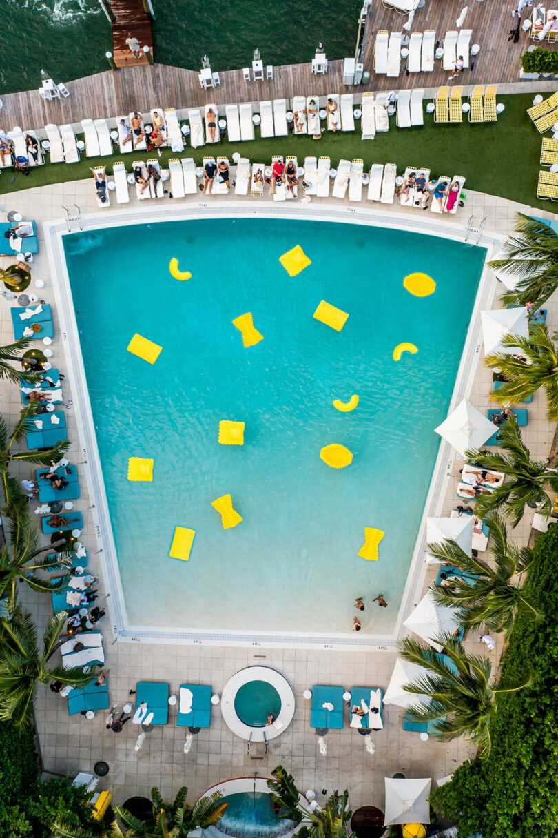 sky view looking down at hotel pool surrounded by people with yellow pasta-like pool floats floating in water