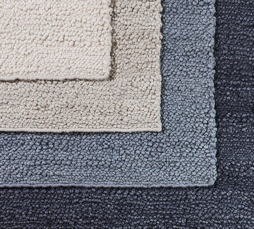 Four rugs layered over one another cropped to show their right hand corner, ranging from a light cream, to dark oat, to light blue to a dark blue rug. The rug's wool and polyester woven texture resembles small pebbles.