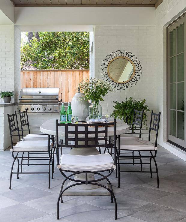 Wrought iron dining chairs sit around an oval marble dining table placed on harlequin pavers beneath a brown plank patio ceiling.