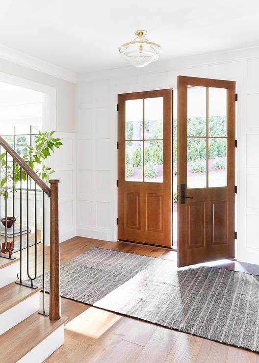 A black and white rug lit by a glass and brass semi flush mount light sits in an entryway in front of brown stained double front doors framed by a board and batten trim.