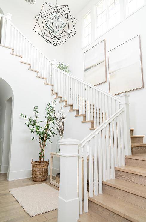 Cottage foyer designed with staggered art on a staircase wall above wood stairs and white spindles. The styled foyer is furnished with a light wood console table and a plant in a basket.