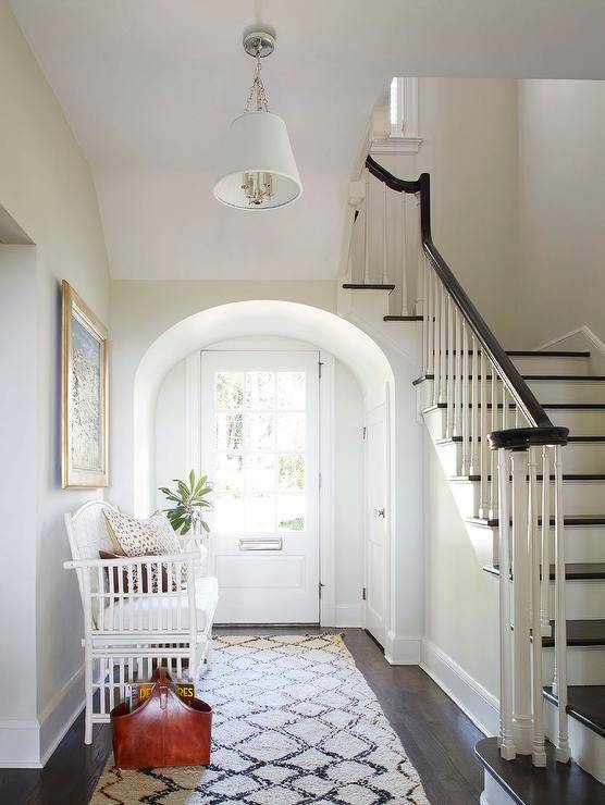 A white front door with glass panels opens to an arched foyer entryway and a Moroccan rug placed beneath a white bamboo settee positioned under a framed art piece.