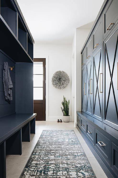 Galley style mudroom featuring navy blue cabinets with shiplap over a built-in bench. X trim adds a modern farmhouse finish to the navy cabinets finished with brass pulls. A gray juju hat decorates the entryway beside a wood door above a snake house plant.