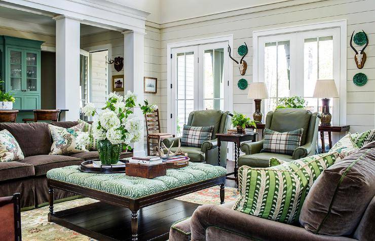 Brown and green living room features a green tufted ottoman coffee table placed on a gren rug between facing brown velvet sofas accented with green, brown, and blue pillows. Green leather club chairs sit side-by-side topped with green plaid pillows.