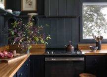 Black kitchen cabinets adorned with black knobs are accented with a butcher block countertop fixed against black shiplap trim and a beneath black shelf that wraps around to a black shiplap range hood. The range hood is mounted over a stainless steel oven range.