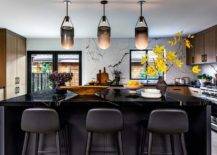 Black center island lit by modern black and white pendants is topped with a black marble countertop paired with black leather stools. Modern brown oak perimeter cabinets are adroned with black pulls and mounted against a white and black marble slab backsplash.