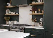 Black and white kitchen features blond floating shelves, a black hood over a white marble slab backsplash and black cabinets with brass pulls.