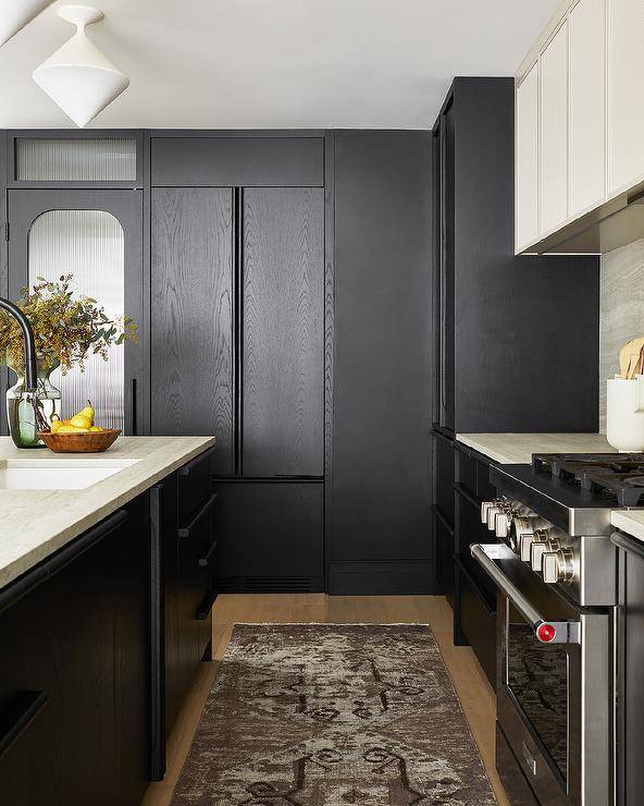 Modern black and ivory kitchen boasts a faded black runner placed between a black oak island topped with an ivory marble countertop lit by white diamond pendant lights and a stainless steel oven range. The stove sits between black cabinets donning an ivory marble countertop and beneath an ivory wood paneled range hood.