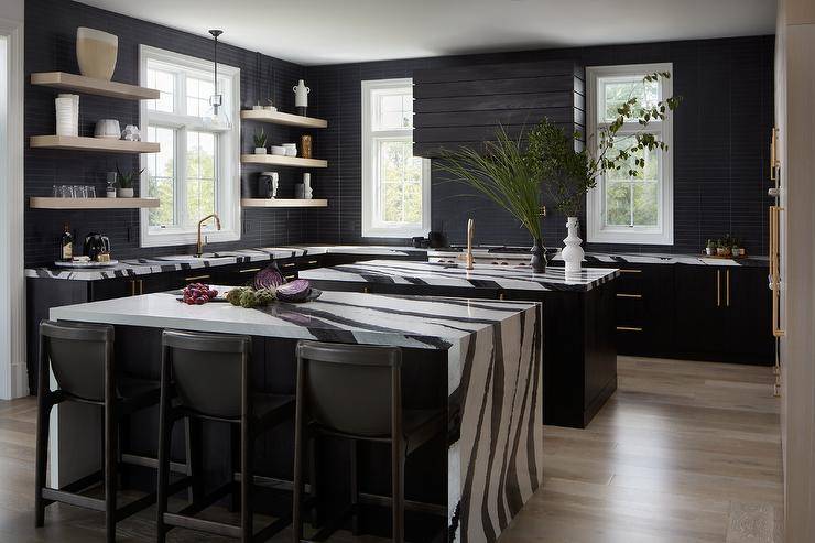 Modern spacious kitchen features a black and white kitchen islands with black leather stools, a black hood over cooktop and light brown floating shelves on black tiles.