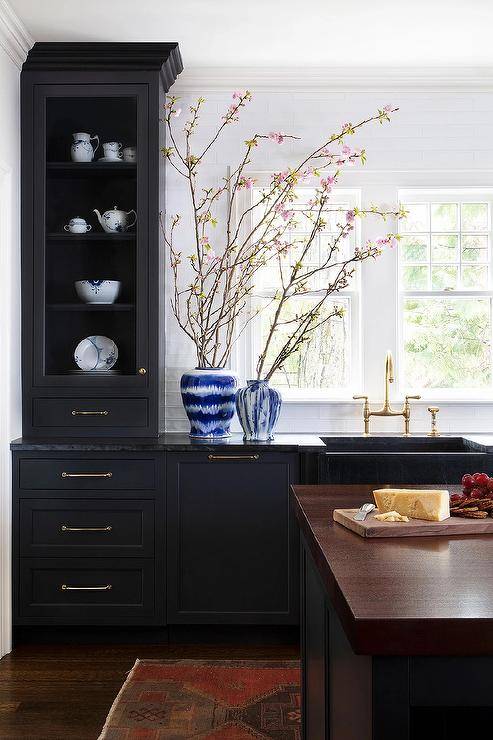 Gorgeous black kitchen features black shaker cabinets accented with brass pulls and holding a black marble apron sink with a brass deck mount faucet beneath a row of windows. The faucet is fixed to a black leathered marble countertop, while white and blue flower vases sit beside a glass front china cabinet.