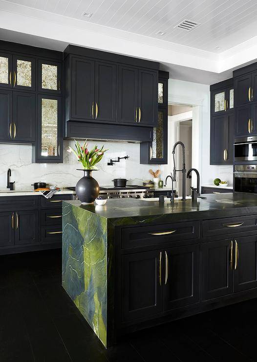 Contemporary kitchen features a green and black marble waterfall island with a matte black pull out kitchen faucet, and antiqued mirrored black cabinets with modern brass pulls topped with ivory and black marble.