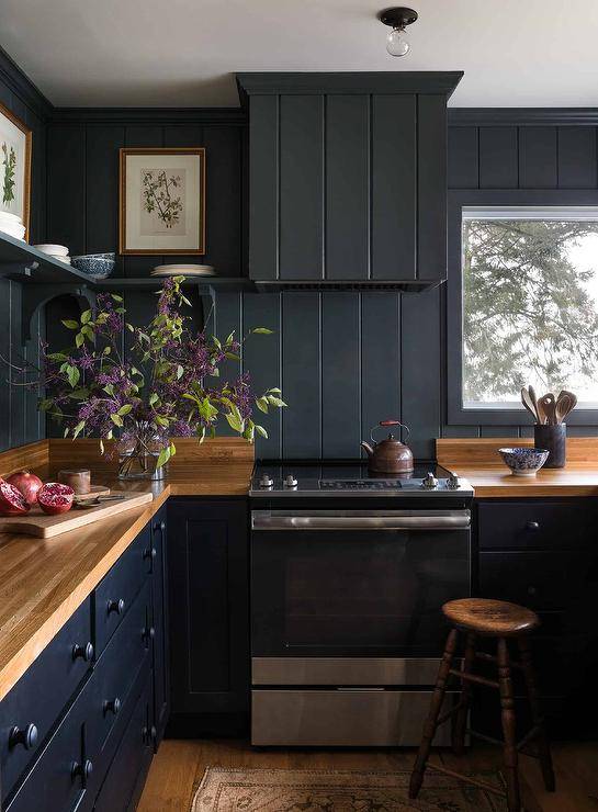 Black kitchen cabinets adorned with black knobs are accented with a butcher block countertop fixed against black shiplap trim and a beneath black shelf that wraps around to a black shiplap range hood. The range hood is mounted over a stainless steel oven range.