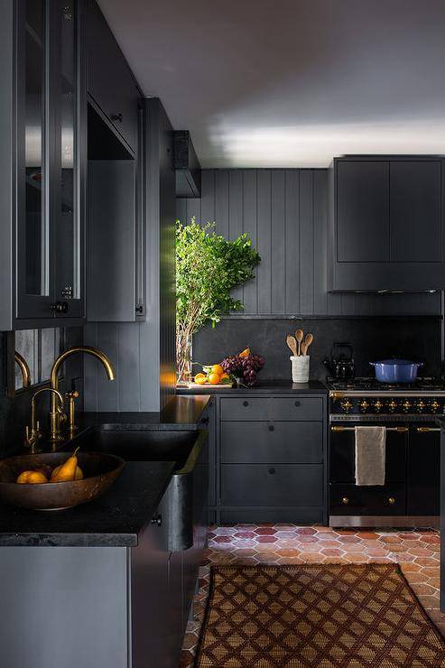 Black kitchen design features black plank cabinets with a black range hood over a black marble slab backsplash and black French stove and red brick hexagon floor tiles.