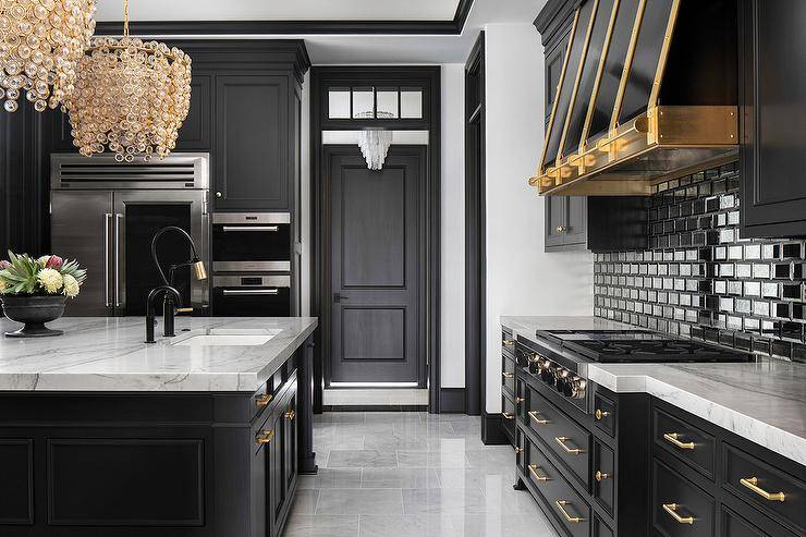 Well-appointed kitchen boasts a gold and black faucet accenting a sink fixed to a gray marble countertop accenting a black island adorned with brass hardware and lit by two gold crystal chandeliers. A black hood with brass straps is mounted against black backsplash tiles over a gas integrated cooktop fixed to black drawers completed with brass hardware.