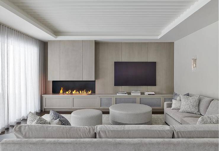 Round gray ottomans sit on a gray rug in front of a gray sectional topped with gray pillows. A gray built-in TV cabinet accneted with mesh doors is fixed against a gray wash plank wall finished with a modern fireplace.