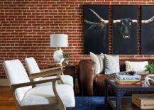 A steer triptych art piece is hung from a red brick wall over a brown leather sofa accented with white and gray pillows in a modern country living room. An industrial rivet coffee table sits in front of the sofa on a bold blue rug and seats two wooden accent chairs with white cushions.