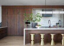 A brown veneer island topped with a marble waterfall countertop is matched with modern brass backless stools and completed with a sink and oil rubbed bronze gooseneck faucet illuminated by a black and white linear chandelier.