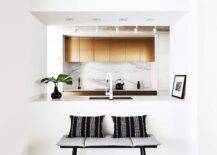 A black bench with gray cushions and black and white striped pillows sits against the back of a kitchen pass through boasting a sink white a white modern gooseneck faucet. Gold cabinets are mounted against a marble slab backsplash over an induction cooktop.