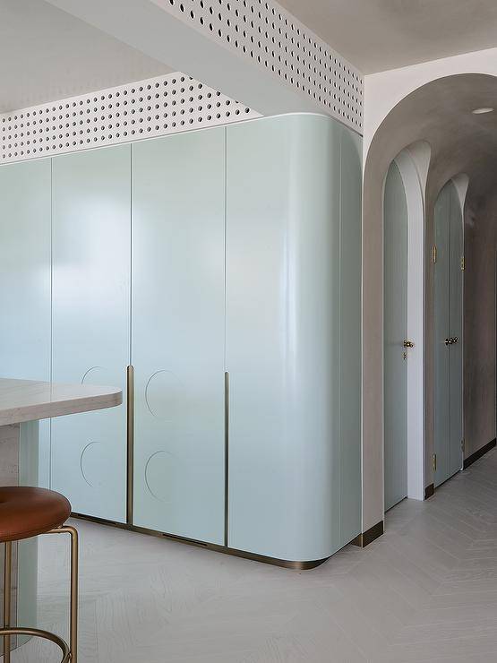 Modern kitchen features light blue cabinetry with brass trim, an island with a brown leather stool and an arched doorway.