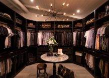 Chic men's large walk-in custom closet features Louis Vuitton luggage under a round marble accent table, illuminated by a sputnik chandelier.
