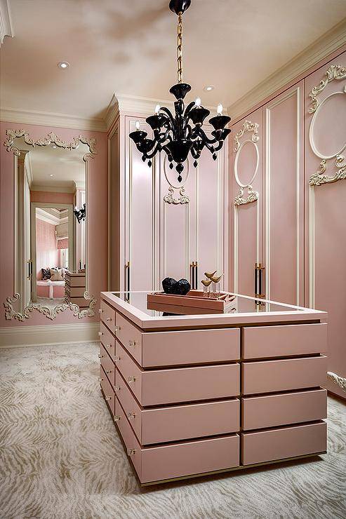 Spacious French pink closet features a pink island illuminated by a black French chandelier and pink cabinets adorned with cream ornate trim.