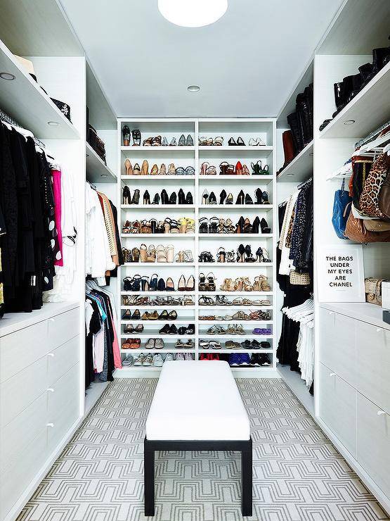 Custom walk in closet features floor to ceiling shoe shelves, built in dressers and a black and white bench on black and white geometric carpeting lit by custom lights.